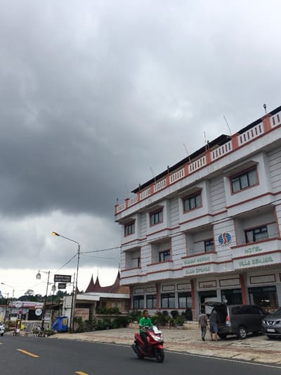 This image depicts a street scene in Palayan, Philippines. The sky is heavily overcast with dark gray clouds.
On the right side of the image, a four-story building with white walls and red trim is prominent. The building features multiple dark-framed windows across its facade. The top floor has an open balcony area with white balustrades and red accents, from which several curved metallic rods extend upwards. Signs on the ground floor are visible, one reading "RUMAH MAKAN ALAM CIPANAS" (restaurant) and another "HOTEL VILLA SENJAYA," both in Indonesian. A logo with an 'S' and wave-like elements is also present on the facade.
In front of the building, on the right side of the road, a dark gray minivan is parked with its rear hatch open. Two adult males are standing near the rear of the minivan; one in a lighter shirt and dark pants faces away from the camera, while another in a darker shirt and shorts is bent over near the open hatch, possibly interacting with cargo. A dark SUV is parked immediately behind the minivan.
In the foreground, a male wearing a bright green t-shirt and dark pants rides a red scooter-style motorcycle from left to right on the asphalt road. Double solid yellow lines are visible on the road surface.
To the left of the main building, a street lamp with a yellow-orange light fixture extends over the road. Beneath it, a black sign with white lettering reads "BUIBUR AYAM NASI PAKET" (Chicken Porridge Rice Package), also in Indonesian. Further left, a structure with a distinctive traditional roof, characterized by upward-curving gables, is partially visible behind trees and additional commercial signs. Various green plants and shrubs line the roadside and building fronts. In the far left background, more distant commercial buildings are visible.