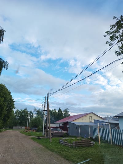 This image depicts a semi-rural residential scene in Malaya Purga, Russia. The sky is partially cloudy, featuring a light blue expanse with scattered white and grey cumulus clouds. Multiple black electrical or communication wires traverse the upper half of the frame, supported by utility poles.

In the foreground, a gravel and dirt road, showing tire tracks, extends from the lower left, curving slightly to the left in the mid-distance. Green grass and weeds line the roadside, and two raised garden beds constructed from corrugated metal are visible in the immediate foreground, containing various plants, including purple irises near a fence.

To the left of the road, dense green deciduous trees form a prominent treeline. A darker vehicle is parked on the grass near a stack of cut firewood. Another utility pole is partially visible among the trees further down the road.

On the right side of the road, a tall wooden utility pole with a metal bracket structure stands prominently, supporting numerous wires. A faint wisp of smoke or steam is observable near the top of this pole. Behind the pole, a corrugated metal shed is positioned in front of a yellow-sided house featuring a red gabled roof and a distinct boarded-up opening on its upper story. A light brown sedan is parked on the grass between the shed and the house. A vertical slat fence, painted with alternating blue and white stripes, partially borders the property in the foreground right. A portion of another house with a grey roof is visible adjacent to the yellow house. No people are visible in the scene. Slight lens anomalies are present in the upper left and lower center areas of the image.