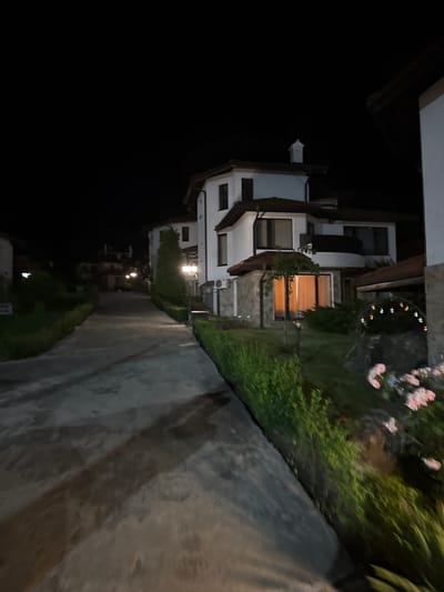 This image depicts a nighttime scene in a residential area within the city of Kosharitsa, Bulgaria. A dark, concrete or paved road ascends gently towards the left, flanked by houses and greenery. On the right side, a prominent multi-story house with white facades and brown tiled roofs features windows, a balcony, and stone cladding on its lower section. Warm light emanates from a ground-floor window of this house. An outdoor air conditioning unit is visible on its left side. In the garden area adjacent to this house, a decorative lamppost illuminates the foreground, revealing green hedges, grass, and pink blooming flowers. Multi-colored string lights are strung over an archway or structure further into the garden. On the left side of the road, more white-walled houses with brown roofs are visible further up the incline, one featuring a white chimney. The dark sky is clear. No individuals or active subjects are present in the scene.