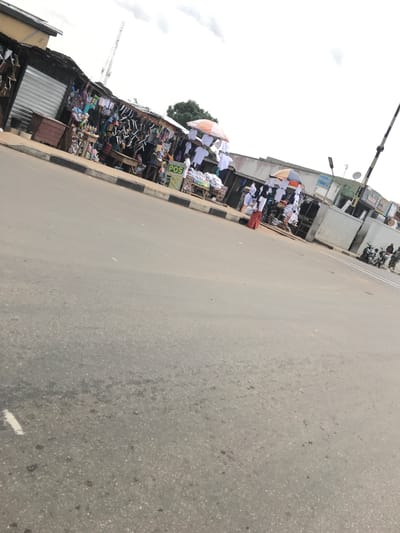 The image depicts a bustling street market scene in Mandate Estate, Ilorin, Nigeria. A wide, grey asphalt road occupies the right side and foreground, showing some texture and faint markings, including a white line at the bottom right. This road is bordered on the left by a black and white striped curb, separating it from the active market area.
Along the left side of the street, numerous makeshift market stalls are visible. These structures are constructed from wood and corrugated iron sheets, creating shaded areas for vendors and their goods. A wide variety of merchandise is on display, including rows of white shirts or garments hanging neatly, some under large umbrellas (one light blue and orange, another white). There's a prominent display of shoes, specifically black and white sneakers, stacked on shelves. Other stalls feature household items, plastic goods, and general merchandise, indicating a diverse array of products available for sale.
Several individuals are present within the market, appearing to be both vendors and shoppers, engaging in daily commercial activities. Some are standing under the umbrellas, while others move between the stalls. Further down the street, two motorcycles are parked on the side, with a few people standing nearby.
The background reveals concrete buildings, some appearing weathered, suggesting established businesses or residences behind the market. The sky is overcast with grey clouds, indicating a cloudy daytime setting. A tall metal structure, possibly a telecommunications mast or a construction crane, is visible in the far-left background. Streetlights and utility poles are also part of the urban infrastructure.
Visible text includes a distinct green sign with "POS" (Point of Sale) written in white letters, affixed to one of the stalls. Another partially obscured blue sign appears to have text resembling "ORIENT." The overall scene captures the vibrant atmosphere of an active local market.
