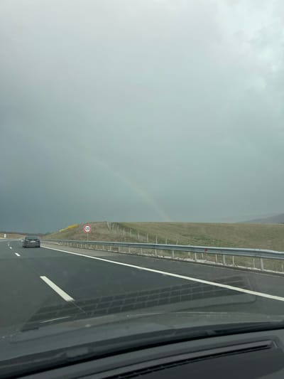 The image shows a highway in Buhovtsi, Bulgaria, possibly taken from inside a moving vehicle. A gray sedan is seen traveling on the road. A metal guardrail runs along the right side of the highway with a speed limit sign of 120. In the distance, a faint rainbow is visible in the sky above the adjacent field. The weather appears overcast.