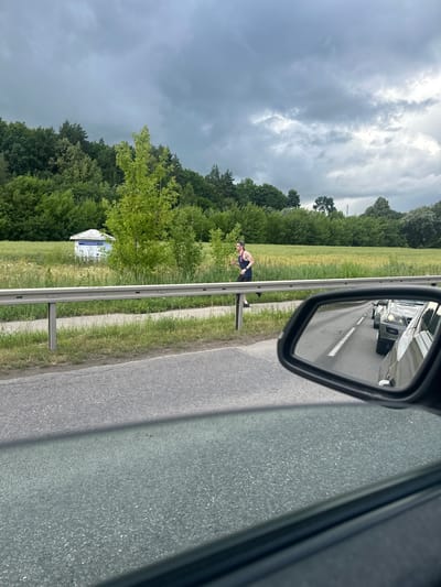 The image shows a woman jogging on a paved path next to a highway. She is wearing a dark tank top and dark shorts, and a baseball cap. She is holding a water bottle in her right hand. The path is separated from the highway by a metal guardrail. In the background, there is a field of green grass and trees. The sky is cloudy and grey. A small white building with a light blue roof is visible in the field to the left of the jogger. The image is taken from inside a car, reflected in the side mirror. The mirror shows a line of cars on the highway. The location is the vicinity of Smiltynai I, Lithuania.