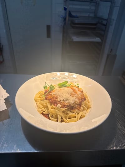 A white plate of fettuccine pasta with bolognese sauce and grated parmesan cheese is displayed. The pasta is topped with a rich red sauce and a generous sprinkle of cheese. A few sprigs of fresh green herbs add a touch of color. The plate is set on a dark gray surface with a metallic sheen, possibly a countertop or table, and is illuminated by soft lighting. In the background, out of focus, is a kitchen or service area with stainless steel appliances and tiled walls. The overall scene suggests a prepared meal ready to be served or enjoyed. There is no visible text. The context indicates this is in Rivesaltes, France.