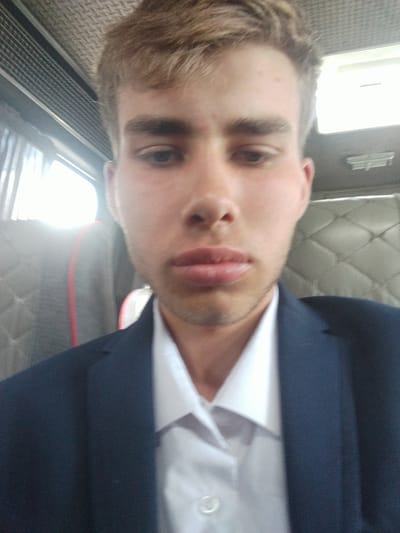A young Caucasian male with light brown hair is captured in a close-up, selfie-style shot. He is wearing a dark blue suit jacket over a white collared shirt. The background appears to be the interior of a vehicle, possibly a bus, with textured grey seating visible. The lighting suggests an indoor or shaded environment, with a bright light source to the left of the frame. The man's face is partially obscured by the camera angle, with his lips and lower nose prominently featured. There is no discernible context regarding his location within the city of Кызыл-Кыргызстан, Kyrgyzstan, or any specific activity he is engaged in.