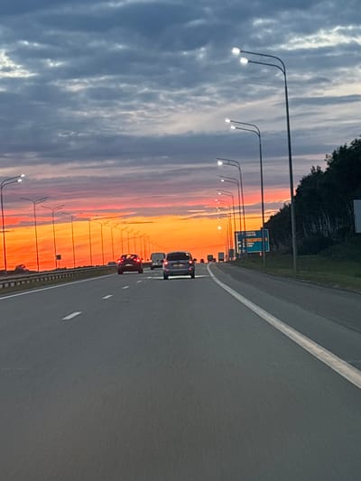 The media file is a vertical, slightly tilted, wide-angle photograph taken from the perspective of a moving vehicle on a highway. The scene is dominated by a dramatic sunset sky, transitioning from deep blue and grey clouds on the left to vibrant orange and pink hues towards the right. A line of streetlights with illuminated bulbs stretches along the left side of the highway, silhouetted against the colorful sky.
The highway itself is paved and appears to have multiple lanes, with white lane markings visible. Several vehicles are traveling on the highway, including cars and what appears to be a van or larger vehicle. The streetlights suggest this is a well-maintained road, possibly an expressway or a major highway.
On the right side of the highway, a line of trees forms a dark backdrop against the sunset. A blue road sign with white text is visible in the distance, indicating directions and distances. Though the text is partially obscured and difficult to read clearly due to the distance and angle, some Cyrillic letters can be discerned, suggesting a location in Russia.
The time of day is clearly sunset, indicated by the intense colors in the sky. The weather appears clear enough to allow for a vibrant sunset. The overall mood is serene and picturesque, capturing the beauty of a sunset on a highway journey. The presence of vehicles suggests a normal flow of traffic. There is no clear indication of people within the frame. The location context provided, Bogorodskoe, Russia, aligns with the visible Cyrillic text on the road sign.