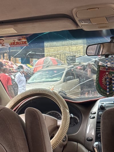 The image, taken from inside a vehicle, shows a cracked windshield obscuring a street scene in Orji, Nigeria. Through the damage, several people are visible, some standing and others near or inside vehicles. One person in the foreground, inside the car, has their arms resting on the steering wheel. Outside the car, there are at least two individuals dressed in casual attire, with one wearing a red t-shirt and the other a white shirt. Further in the background, more people and vehicles are present, with umbrellas providing shade in what appears to be a marketplace or busy street. A political sticker with the words "LABOUR PARTY" is hanging from the rearview mirror. The overall environment suggests a busy urban setting, likely during the daytime.