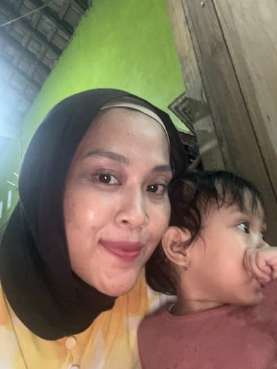 In an indoor setting in Wedarijaksa, Indonesia, a woman wearing a black hijab and a yellow and white patterned shirt is taking a selfie. She is positioned to the left of the frame, with her face turned towards the camera, smiling. Next to her, to the right, a young child with dark hair is visible. The child is wearing a rust-colored shirt and has their hand near their mouth. The background features a bright green wall and a wooden structure.