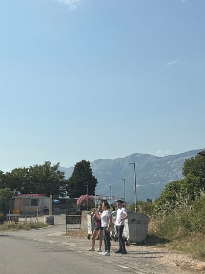 The image depicts a group of three young people standing by a road in Blizikuće, Montenegro. Two are wearing white t-shirts and dark bottoms, while one is in a white t-shirt and burgundy shorts. They appear to be conversing or posing for the photo. In the background, a large mountain range rises majestically against a clear blue sky, suggesting a bright, sunny day.
To their right, a gray dumpster sits beside the road. Further down the road, several signs are visible, including one that reads "MARKET" in red lettering on a small building. Other signs indicate "OPEN", "RESTAURANT", "MEAT FISH", and "ROOMS", suggesting this is a commercial area with various amenities for travelers. Trees and dense vegetation line the roadside and the slopes of the mountain, adding to the natural beauty of the scene.
The overall impression is one of a casual, outdoor encounter in a scenic, mountainous region of Montenegro. The time of day appears to be midday, given the bright sunlight and the shadows cast. The mood seems relaxed and informal.