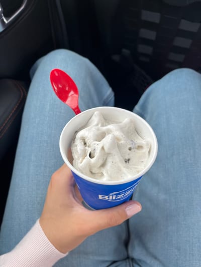 The image shows a person holding a Dairy Queen Blizzard in a car. The Blizzard is Cookies and Cream. A red plastic spoon rests inside the cup. The person wears light blue jeans and a light pink sweater. The interior of the car is visible. The location is the city of Johnson City, United States of America.