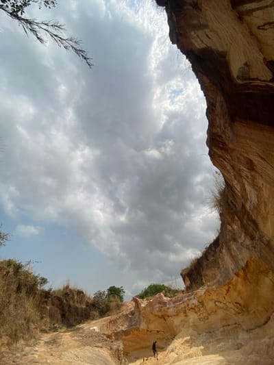This forensic image depicts an open excavation site or natural gully within Amangwu, Nigeria. The scene is dominated by a tall, stratified cliff face on the right, composed of light brown, orange, and reddish-brown earth layers, with sparse dry vegetation clinging to its edges. The ground slopes from left to center, featuring a dirt path and areas covered in dry, golden-brown grasses, with green trees visible in the distant background. A lower, lighter-colored basin of yellowish-white soil occupies the bottom-center and right foreground. A lone individual, dressed in dark clothing, stands within this lower basin near the base of the prominent cliff. The sky above is largely overcast with dense grey clouds, though patches of lighter blue sky are discernible. A bare tree branch extends into the upper-left corner of the frame.