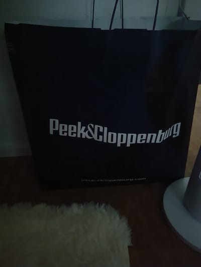 The image shows a dark blue shopping bag with white lettering. The bag is standing upright, and the front of the bag is visible. The text on the bag reads "Peek & Cloppenburg" in large, stylized letters. Below this, in smaller text, it says "peek-cloppenburg.com". The bag appears to be made of paper and has two dark straps at the top.

The bag is placed on a wooden floor. To the left of the bag, there is a fluffy, light-colored rug or carpet. To the right of the bag, a portion of a white, circular object, possibly a lamp base or a stand, is visible. The background behind the bag is a light-colored wall.

The lighting in the image is dim, suggesting it might be evening or nighttime, or the room is not brightly lit. There are no people visible in the image. The overall impression is that of a retail shopping bag, possibly after a purchase has been made. The location context provided is Germersheim, Germany, which is consistent with Peek & Cloppenburg being a German clothing retailer.
