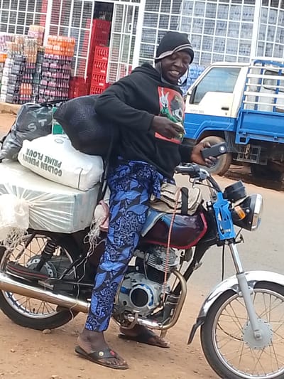 A man in Iseyin, Nigeria, sits astride a motorcycle laden with goods. He wears a black beanie, a black hoodie with a graphic print, and blue patterned trousers. His right hand holds a black object, possibly a phone, while his left hand is held out, fingers spread in a gesture. The motorcycle is dark-colored with red accents, and it carries a large white sack labeled "GOLDEN PENNY PRIME FLOUR" and other wrapped items. In the background, a blue and white truck is parked, and stacks of crates, some red and some containing bottled drinks, are visible outside a building with a white, grid-like facade. The ground is dusty.
