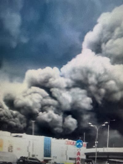 The image captures a chaotic scene dominated by thick, dark smoke billowing from what appears to be a building or industrial structure. The smoke is voluminous and turbulent, obscuring much of the background and creating a dramatic, almost apocalyptic atmosphere. To the right of the frame, a tall, white structure is visible, potentially a building or part of a larger industrial complex. This structure has several horizontal bands of color, including a prominent blue band and what looks like red and white striped tape, possibly indicating a hazard zone or caution tape. A circular blue sign with a white symbol, possibly a traffic or warning sign, is also visible on this structure.

The scene is characterized by an intense, dramatic visual. The presence of smoke strongly suggests an emergency event such as a fire or explosion. The surrounding environment appears to be urban or industrial, given the presence of the building and what might be street furniture or infrastructure to the right. There are no clear signs of people in the immediate vicinity of the smoke, but the context of an emergency event implies the potential presence of emergency responders or affected individuals. The lighting suggests it could be daytime, as the smoke is illuminated from above, but the overall darkness of the smoke makes it difficult to ascertain the exact time. The location is identified as Sof'ino, Russia. There is no discernible emotion visible beyond the inherent drama of the event itself.