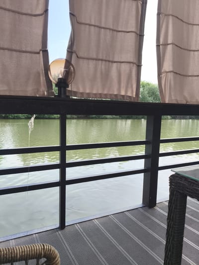 This image depicts a tranquil scene from a covered outdoor seating area overlooking a calm body of water, likely a lake or river, in Komsomolskoe, Kyrgyzstan. The foreground shows a dark, slatted wooden deck with a portion of a wicker chair visible on the left and the woven leg of a table on the right, suggesting a dining or relaxation setting. A black metal railing separates the deck from the water, with a single, detached strand of white string hanging from it.

Above the railing, several panels of light brown fabric hang as a curtain, obscuring parts of the sky and a single, round, amber-colored lamp with a visible filament. Through the gaps in the curtains, a bright blue sky is visible, hinting at fair weather. Beyond the railing and the water, lush green trees line the far bank of the water, suggesting a natural, possibly forested, environment. The water itself is a murky green, with subtle ripples on its surface reflecting the light.

The lighting suggests it is daytime, likely late morning or early afternoon, given the bright sky and the clarity of the scene. There is no discernible activity taking place, and the overall mood is peaceful and serene. The visual elements combine to create a sense of quiet repose and natural beauty, enhanced by the subtle textures of the wood, wicker, fabric, and water. No text is visible in the image.