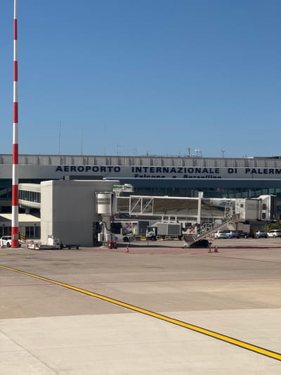 The image depicts the Falcone–Borsellino Airport (Aeroporto Internazionale di Palermo Falcone e Borsellino) in Cinisi, Italy. The airport terminal building is visible, with the airport's name prominently displayed vertically in blue lettering. Ground service equipment, including jet bridges and vehicles, is positioned adjacent to the terminal. A large expanse of concrete apron or taxiway extends to the right, marked with yellow lines. In the foreground, a red and white striped barrier or pole is partially visible, indicating a restricted area. The sky is clear and blue. There are no people visible in the image.