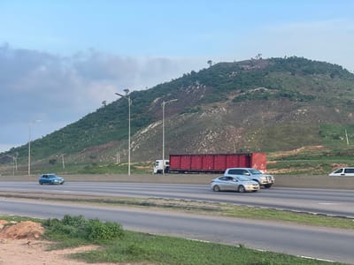 The image captures a highway scene in Immigration Quarters, Nigeria, under a partly cloudy sky that suggests it is either early morning or late afternoon. The foreground shows a paved road with grassy verges and some loose soil and rocks on the left. Several vehicles are moving on the highway.
In the middle ground, a light blue car is seen on the leftmost lane. Further down the road, a silver sedan is in motion, followed by a silver SUV and a large white truck with a red cargo container filled with what appear to be red crates. A white van is visible on the far right lane. The highway is bordered by a concrete barrier.
The background features a prominent, large, verdant hill with patches of exposed earth and rocks, characteristic of a mountainous or hilly landscape. Several tall streetlights are positioned along the highway. The overall atmosphere is calm, with the activity focused on the movement of traffic along the road against the backdrop of the natural landscape. No discernable text is visible in the image.