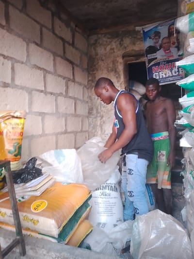 The image shows two men inside a small, somewhat dim room or storage area in Mgbuoshimini, Nigeria. The walls are constructed from rough concrete blocks, suggesting a utilitarian or unfinished space.

In the center, a man wearing a black sleeveless shirt with white accents and light blue jeans with a repeating "Chanel" logo pattern is bent over, handling a large clear plastic bag or a white sack, possibly filled with grains. To his right and slightly behind him, a second man with a bare torso and green and orange patterned shorts stands, looking in the direction of the first man's activity. Both men appear focused, with neutral expressions.

The room is filled with numerous bags, mostly large sacks, indicating it's a place for storing or selling bulk goods like rice. To the left, a yellow bag is partially visible with text such as "NBAG TO WIN CASH," "EVERYS," and "50" (likely indicating quantity or weight). Below it are stacked items resembling books or notebooks. A prominent white sack in the foreground clearly displays "Parboiled ARMTAIN Rice," "Grown in Nigeria to Feed Africa," and "50kg."

On the back wall, to the right, a poster is affixed, featuring images of individuals and advertising "EXPERIENCE The HOUR GRACE." Below this poster, a green bag can be seen with "MADE IN EGYPT" printed on it. The scene suggests a small-scale commercial operation, likely a grain or foodstuff shop, during daytime hours, though the interior lighting is somewhat subdued.