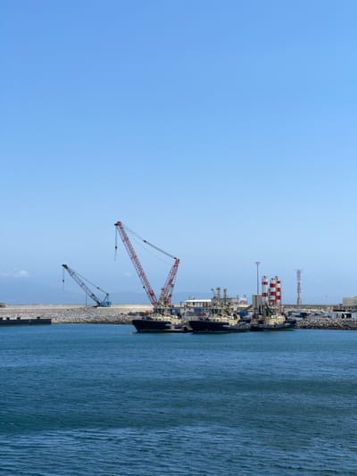 The image shows a maritime industrial scene in the port of Ksar al Majaz, Morocco, under a clear blue sky. In the foreground, the blue water of the harbor is calm with gentle ripples. In the middle ground, a stone breakwater forms a barrier between the water and the industrial area. Several tugboats are docked along the breakwater. Two large cranes are visible on the breakwater: a tall red lattice boom crane on the right and a blue and black lattice boom crane further to the left. Behind the cranes, there are industrial buildings and structures, including two tall silos with red and white checkered patterns and a radio tower. In the distance, hazy mountains form the backdrop. The overall scene is one of port activity and industrial infrastructure.