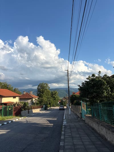 This is a photograph of a street in the town of Anton, Bulgaria, under a partly cloudy blue sky. The road, paved with asphalt, runs diagonally from the lower left to the upper right of the frame, with a gray tiled sidewalk running parallel to it on the right. The sidewalk is bordered by a low stone wall topped with a green, ornate metal fence.
To the left of the road, houses with red-tiled roofs are visible, separated by green and brown fences. Trees and vegetation are present in front of and between the houses.
A wooden utility pole with numerous black wires stretches across the upper right portion of the image, its wires fanning out against the sky. In the distance, rolling hills covered in green foliage and trees form the backdrop. A white car is parked on the right side of the road, facing away from the camera. The scene is illuminated by natural daylight, casting shadows on the road and sidewalk.