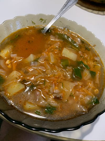 The image shows a close-up of a bowl of soup, likely a hearty stew, presented in a decorative scalloped bowl. The soup is a rich, orangey-brown broth filled with visible ingredients such as chunks of potato, shredded cabbage, and chopped green vegetables, possibly green onions or peppers. Finely chopped dill is scattered throughout, adding a fresh, herbal element. A silver spoon rests in the left side of the bowl, partially submerged in the broth, suggesting the soup is being eaten or is about to be. The lighting appears to be indoor, casting subtle shadows and highlighting the oily sheen on the surface of the soup, indicating it is hot. The setting is likely a home environment, given the simple white tablecloth beneath the bowl. The overall impression is one of a warm, comforting meal. There is no visible text in the image.