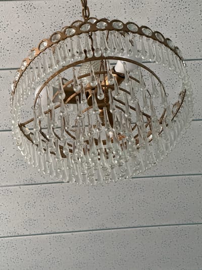 An ornate, circular chandelier hangs from a chain, its brass frame adorned with numerous clear glass teardrop prisms. The chandelier is suspended against a backdrop of textured wallpaper featuring a repeating pattern of stylized floral or starburst motifs in a light gray color. The wallpaper is divided into horizontal sections by thin, lighter-colored bands. The image is taken from a low angle looking upwards at the chandelier. There are no people or animals visible. The overall impression is one of an indoor setting, likely a room with decorative ceiling treatment. The location in Jambay, Uzbekistan is not visually evident from the image itself.