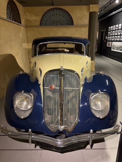 The image, oriented vertically, captures a vintage two-tone car on display, likely within a museum or exhibition hall in Wassenaar, Netherlands. The car's body features a distinct dark blue on its upper half and a creamy light beige on its lower half, including the fenders. The front of the vehicle is prominent, showcasing a large, chrome-plated grille composed of numerous horizontal bars. A circular red emblem, possibly a brand insignia with stylized letters, is centered on the grille. Flanking the grille are two large, circular headlamps, each with a chrome surround, embedded in the dark blue bodywork. A shiny, robust chrome bumper extends across the very front of the car.
The car is situated indoors, with warm-toned, possibly plaster or concrete walls forming the immediate background. On the left side of the frame, two decorative, circular metal grilles are visible, seemingly integrated into the floor or wall. In the upper background, a dark wall or ceiling area features a precise grid display of multiple framed black-and-white photographs or historical documents, characteristic of a museum exhibit. The setting is illuminated by artificial indoor lighting. No people are visible, focusing attention solely on the meticulously presented classic automobile. The overall impression is one of historical preservation and artistic display.