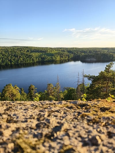 The image shows a serene landscape with a body of water, likely a lake, surrounded by dense green forest under a clear blue sky with scattered white clouds. In the foreground, a rough, rocky surface with patches of moss is visible, blurred due to a shallow depth of field. The lighting suggests it is either early morning or late afternoon, with the sun casting a warm glow on the scene. The location is identified as Skriketorp, Sweden. No people or animals are present in the image.