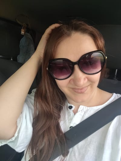 In the city of Tselina, Russia, a woman with long brown hair, wearing sunglasses and a white shirt, is captured in a selfie taken from inside a vehicle. She is placing her hand on her head, and a seatbelt is visible across her chest. The background is dark, suggesting the interior of a car. A small grey object with lace detailing is hanging from the car's interior, possibly an air freshener.