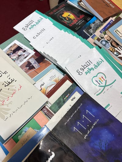 The image displays a close-up, high-angle view of numerous books stacked and fanned out on a surface, presumed to be within a building in Al-Ghizlaniya, Syria. The underlying surface appears to be red, possibly a tablecloth or carpet. Several identical white booklets titled "التطوع أخلاق وقيم" (Volunteering: Ethics and Values) are prominently visible, featuring a logo of four stylized, multi-colored hands forming a heart shape with intertwining lines. Other book titles include "THE SIGN OF FOUR" by Sir Arthur Conan Doyle, alongside its Arabic translation "علامة الأربعة," and a dark-covered book with large white numerals "11:11" against a starry night sky with a bare tree silhouette and Arabic text below. Another book partially visible has an Arabic title translating to "O Awakening... to free the soul from oppression..." Various other books with Arabic text and diverse cover art, some showing historical figures or photographic prints, are also present. No individuals are depicted, and no explicit actions or interactions are occurring. The scene is static, focused entirely on the arrangement of books.