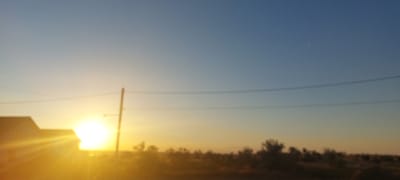 The image depicts a sunset scene with a bright sun on the left side, casting a strong glare. A silhouetted building with a pointed roof is visible to the left of the sun. A utility pole with wires stretches horizontally across the frame, separating the sky from the lower portion of the image. The lower part of the image shows a blurred landscape with trees and vegetation, suggesting an outdoor environment. The sky transitions from a warm orange hue near the horizon to a clear blue at the top. The scene is set in Korsuntsi, Ukraine.