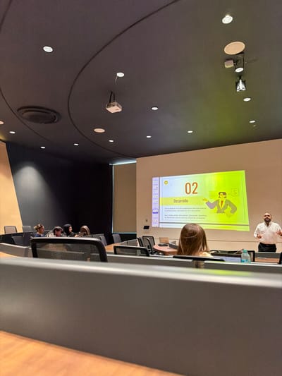 A modern tiered lecture hall or conference room is depicted in Fraccionamiento Los Heroes Queretaro, Mexico. The room features a dark gray, curved ceiling with numerous recessed circular lights and a ceiling-mounted white projector. Walls are a combination of light beige and dark gray. A large white screen on the front wall displays a presentation slide with a bright yellow-green background, showing the number "02," the Spanish word "Desarrollo" (Development), and an illustration of a man speaking with a dollar sign. A male presenter with a beard, wearing a light-colored collared shirt and dark pants, stands to the right of the screen, facing the audience with his hands slightly open. In the foreground, the back of a person's head with light brown hair is visible, seated at a dark gray desk. Further back and to the left, approximately five individuals are seated in black mesh-backed chairs at dark gray desks, facing the presenter. A clear plastic water bottle is visible on a desk in the foreground.