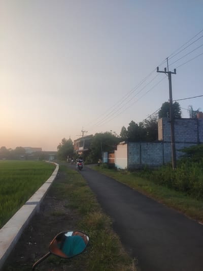 The image depicts a rural road in Sidokepung, Indonesia, during what appears to be sunrise or sunset, indicated by the soft, warm light and gradient sky. A narrow asphalt road curves to the right, bordered by a green rice field on the left and overgrown vegetation and buildings on the right.

On the road, two individuals are seen riding motorbikes. The motorbike in the foreground is closer to the viewer, and a person wearing a turquoise shirt is visible in the reflection of its side mirror. The second motorbike, with two people on it, is further down the road, heading away from the viewer.

The environment includes a concrete barrier along the edge of the rice field, a utility pole with multiple wires, and several buildings of varying construction, including one with a grey brick wall. Lush green trees and bushes are scattered throughout the scene, with some appearing to be in front of the buildings. The overall atmosphere is tranquil and suggestive of daily life in a rural Indonesian village.