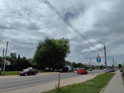 The image depicts a street scene in Diveyevo, Russia, under a cloudy sky. A paved road runs horizontally across the foreground, with a sidewalk on the right side. Several cars are parked along the road and on the grass verge. In the distance, a golden-domed building, likely a church, is visible. Power lines stretch across the sky, supported by utility poles. The overall impression is one of a quiet, somewhat overcast day in a provincial town.
