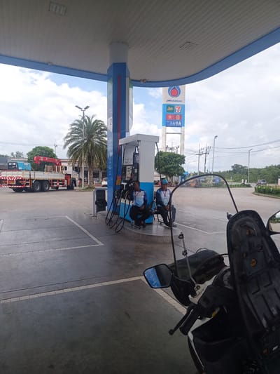 A gas station in Tha Ruea, Thailand, is captured in a full, daylight outdoor shot. The foreground features a parked, white and black motorcycle with a windscreen. Behind it, two men in matching light blue shirts and darker blue pants are seated on a bench alongside a gas pump.

A large, red and white flatbed truck with a red crane sits idle in the background. Palm trees and utility poles are visible, further adding to the environment. A tall, blue and white sign prominently displays the "ptt station" logo, along with a 7-Eleven emblem. The sky is overcast. The gas station features a canopy over the pumps supported by blue and white pillars.