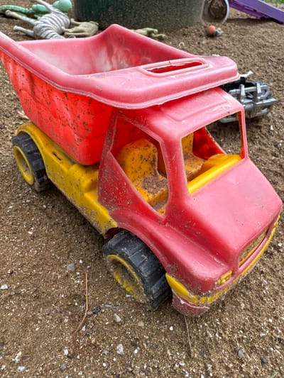A red and yellow toy dump truck sits on a sandy surface, likely a playground or sandpit, in Khazar Raion, Azerbaijan. The truck is partially filled with dirt. In the background, other toys are visible, including a green and white striped toy with a ribbed surface and a small, light brown, worm-like object. The sand contains small pebbles and shells. The lighting suggests an outdoor setting.