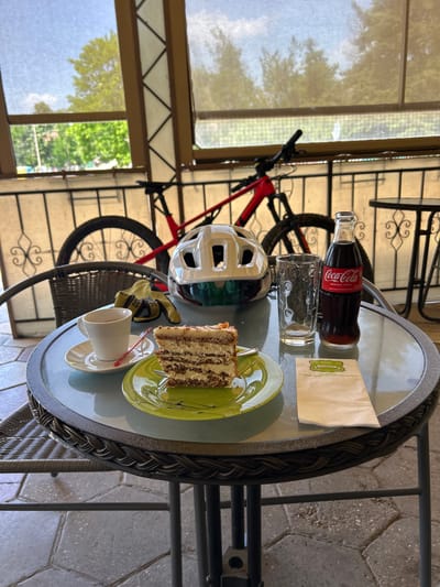 The image depicts a static, outdoor cafe scene in the village of Profesor Ishirkovo, Bulgaria, under daylight conditions. The primary subject is a round glass-top table with a dark metal frame featuring a braided texture around its edge.
On the table, a slice of multi-layered cake with light cream filling and a garnish, possibly nuts or caramel, rests on a light green plate accompanied by a small fork. To its left, a white espresso cup sits on a white saucer with a small red spoon. Further to the right, a clear, textured empty drinking glass stands next to a classic glass bottle of Coca-Cola "Original Taste," which is approximately one-quarter full. A folded white paper napkin, displaying Cyrillic text with a green logo and the inscription "с. Проф. Иширково" (village Prof. Ishirkovo) and a phone number, is placed in front of the Coca-Cola bottle. Also on the table, to the left of the drinks, is a white bicycle helmet with a dark visor, next to a pair of yellow and black cycling gloves.
In the background, a red mountain bicycle is parked, with its front wheel, handlebars, and part of the frame visible on the left, and the rear wheel and part of the frame on the right. The setting appears to be a covered patio or veranda, enclosed by metal railings and support structures. Translucent roll-down blinds are visible in the upper sections of the enclosure, filtering sunlight. Beyond the railing, a lush green outdoor environment with trees and foliage is discernible under a bright, partly cloudy sky. The flooring consists of grey, interlocking paved tiles. Another cafe table with a similar design is partially visible on the far right. No people are present in the scene.