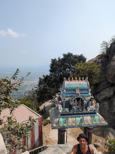 The image depicts a high vantage point overlooking a valley in Kadaladi, India. In the immediate foreground, the upper torso of a man is visible, facing the viewer. He wears a dark tank top, a dark brimmed hat, and exhibits tattoos on his left arm. Green foliage partially obscures the lower left of the frame. Directly behind the man and slightly below him stands an ornate, colorful Hindu temple structure, characterized by a multi-tiered, intricately decorated roof with sculpted figures of deities and animals (including elephants) and topped with golden finials. The structure is supported by four pillars. To the left of the temple structure, a rectangular building with a reddish flat roof and a lower section painted with red and white vertical stripes is partially visible, surrounded by vegetation. A dirt path descends from the temple structure. In the mid-distance on this path, two or three indistinct human figures are present. The background reveals a vast landscape of fields and scattered vegetation in a valley, stretching towards distant hills under a clear, pale blue sky with minimal cloud cover. Large natural rock formations are prominent on the right side of the scene, indicating a mountainous or hilly environment. The scene is illuminated by bright daylight, casting clear shadows.