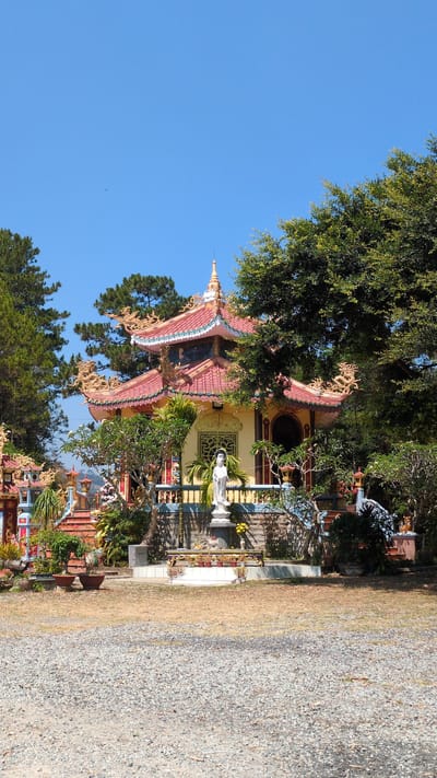 A color, eye-level outdoor shot shows a shrine at the Buddhist Temple in Lâm Sơn, Vietnam, in daylight, and set to the side with a pathway and blue sky.
The Buddhist temple is ornate and multicolored, with layered tiers of red-tiled roofs. Golden dragon statues adorn the edges of the roofs. A single white statue stands in front of the temple. A large tree casts a shadow on the left side of the temple.
The ground next to the temple is covered in gravel and dried grass. The sky is clear and blue.