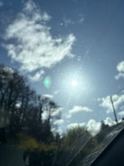 The image depicts an outdoor scene in the city of Ruyton-XI-Towns, United Kingdom, viewed through a transparent, soiled, and scratched surface, likely a vehicle's windshield. The upper and central portions reveal a bright blue sky with scattered white cumulus clouds. A radiant sun is positioned in the upper right, generating a pronounced greenish circular lens flare slightly to its left and a starburst effect around its own brilliant core. The foreground glass surface is marred by numerous small, irregular specks, consistent with dried water droplets or dust, and multiple fine linear abrasions. The mid-ground and background feature out-of-focus dark silhouettes of trees, some appearing bare and deciduous, alongside areas of lighter green foliage, primarily on the left and lower-left. A dark, vertical post, possibly a utility pole, is faintly visible on the far right. The lower right frame shows an indistinct, dark section indicative of a vehicle's interior trim. No human figures or specific interactions are present.