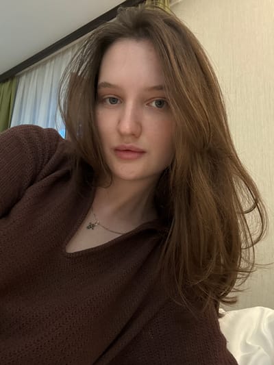 A medium shot captures a young woman from the chest up, facing the camera. She has fair skin, light blue eyes, and long, wavy, brown hair that frames her face and cascades over her shoulder. She is wearing a dark brown, textured sweater with a V-neck opening. A delicate silver necklace with a cross pendant rests on her collarbone. The background is a plain, light beige wall with a section of green and white curtains visible to the left. The lighting is soft and appears to be coming from an indoor source, possibly a lamp or natural light from a window. The overall impression is a casual, indoor portrait. The location is identified as the city of Журавлево (кемеровский), Russia.
