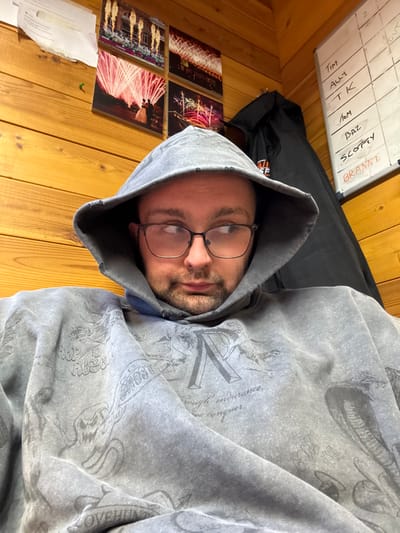 A male individual with light skin, dark-rimmed glasses, and light facial stubble is seated indoors, partially obscured by the hood of a distressed grey hooded sweatshirt adorned with various dark grey graphics, including skulls, text, and a cobra motif. He is looking towards the upper right of the frame. The setting is an indoor room, described as being in Whiteparish, United Kingdom, featuring light brown wooden paneling on the walls. Behind the individual, to the left, four square-framed pictures of fireworks displays are arranged in a 2x2 grid, with a piece of white paper partially visible above them. To the right, a dark jacket hangs on the wall beside a white board. The whiteboard lists names vertically in black marker: "TIM," "ALLY," "TK," "TOM," "BAZ," "SCOTTY," and "BRANNI," with "BRANNI" distinctly written in red marker. The environment is illuminated by artificial light.