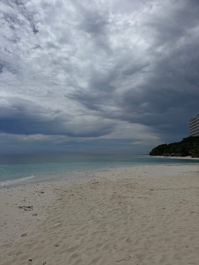The image depicts a beach scene in Ukulhas, Maldives. The sky is filled with layered, textured clouds ranging from white to dark gray. The ocean transitions from a light turquoise near the shore to a deeper blue further out. A strip of beach with light-colored sand shows footprints and scattered debris. In the far distance, a building and some green vegetation are visible.