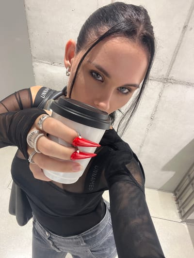 A female individual is depicted in a close-up, angled shot, appearing to be a selfie. She has dark hair, pulled back with strands framing her face, and blue-grey eyes with dark eyeliner. She wears silver hoop earrings. Her attire includes a black sheer mesh long-sleeve top over a black garment with an exposed strap bearing the word "UNNAMED," and light grey denim jeans. Her left hand holds a white disposable coffee cup with a black lid, positioned near her mouth, partially obscuring the lower facial area. The fingers of her left hand are adorned with several silver rings, including a textured ring on the index finger and a coiled ring on the middle finger. Two prominent, sharply pointed, bright red artificial nail extensions, each secured with a silver ring, are visible on her middle and ring fingers. The background consists of light grey, concrete-textured walls with visible seams, indicative of an interior, modern industrial aesthetic. A subtle light source is visible in the upper right. This scene is located in Seongdong-gu, South Korea.