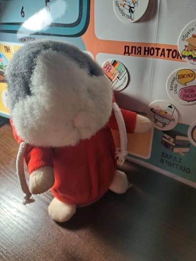 This is a close-up, eye-level shot of a stuffed toy with gray and white fur and a red outfit. The toy is positioned on a dark wooden surface, likely a table. In the background, a colorful, glossy poster is visible. The poster features various text and circular graphics. Prominent on the poster are Russian phrases such as "ДЛЯ НОТАТОК" (for notes) and "ЗАРЯЗ Я ЧИТАЮ" (charge I read). There are also graphics depicting a karateka, flags of different countries (UK, Germany, France), a train, and books. The lighting appears to be artificial, casting highlights on the poster. The image does not contain any elements that would allow for determining its location in Pervomaisk, Ukraine.