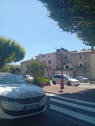 In Viviers, France, a sunlit street scene unfolds. A white Peugeot 508 is parked prominently in the foreground, its license plate reading FZ-934-AK. Across the street, a silver minivan and a silver coupe are also parked. Buildings with tiled roofs and pale facades line the street, some featuring shuttered windows. A red streetlamp stands near the minivan, and a flowering bush with pink blossoms adds a touch of color to the scene. Trees with green foliage cast shadows on the street and sidewalk. A crosswalk with white stripes is visible in the lower portion of the image. The sky is a clear, bright blue.