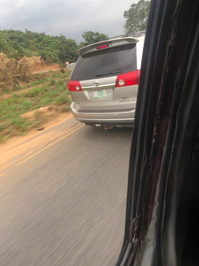 From the perspective of a passenger in a car, a silver Toyota Sienna follows closely behind. The Sienna's rear is visible, featuring black paneling, red taillights, and a Nigerian license plate. The vehicles are traveling on a paved road with double yellow lines, bordered by green vegetation and trees. The sky is overcast. The top portion of the car's interior frame is visible, adding depth to the shot.