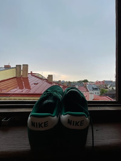 The image shows a pair of green Nike sneakers resting on a wooden surface in the foreground. The sneakers are positioned facing away from the viewer, with the "NIKE" logo clearly visible on the heel of each shoe. In the background, a cityscape is visible through an open window frame, suggesting an urban setting. The cityscape features red-tiled roofs, brick chimneys, and various buildings, indicative of older European architecture. The sky is overcast, suggesting a cloudy day. The overall scene conveys a sense of everyday life, perhaps a moment of relaxation or contemplation with a view of the city. The specific location of Jihlava, Czechia, is not explicitly indicated by any visual cues in the image itself.