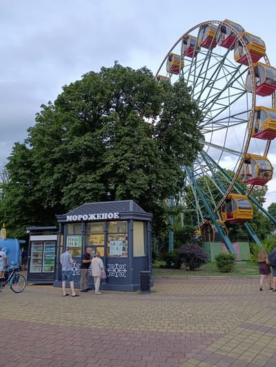 The image depicts an outdoor scene in an amusement park in Kelermesskaya, Russia. Dominating the background is a large Ferris wheel with yellow and red cabins. In the middle ground, a lush green tree stands tall next to a small, dark-colored kiosk with the Russian word "МОРОЖЕНОЕ" (ice cream) illuminated on its facade. Several people are present: two men and a woman are gathered near the kiosk, appearing to be ordering or interacting with staff. To the left of the kiosk is a refrigerated display cabinet filled with drinks, and a bicycle is parked nearby. Further to the right, a couple walks away from the camera. The ground is paved with red and yellow bricks. The sky is overcast.