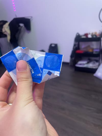 A close-up shot shows a person's thumb and index finger holding a small, crumpled blue and white wrapper. The wrapper is made of a thin, foil-like material and has a checkered pattern with a stylized blue globe and text printed on it. Some of the text appears to be in Cyrillic characters, possibly indicating "САЛФЕТКА" which translates to "napkin" or "wipe". The background is out of focus and appears to be an indoor setting with a wall lit by purple light and a dark wooden floor. There are objects visible on shelves in the background, but they are indistinct. No location in Romanovka, Russia is discernible from the image.