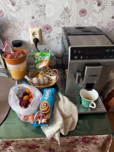 The image depicts a close-up, high-angle view of a kitchen counter in Skopin, Russia. The scene is dominated by a De'Longhi coffee machine, which is actively brewing coffee into a green and white patterned mug. In front of the coffee machine, a folded white waffle-weave towel rests on the counter. To the left of the coffee machine, a plastic bag filled with cherries and apricots sits on the counter. Adjacent to the fruit bag is a blue package of "Rollet" biscuits. Further left, a woven basket contains garlic and a wedge of lemon. Behind the basket, a large orange bowl holds what appear to be packets of sweetener or tea. To the left of the orange bowl is a transparent plastic container filled with a yellow liquid, possibly honey or oil. A black electrical outlet with a black plug is visible on a patterned wallpapered wall in the background. The counter itself is a dark green surface, and the foreground is partially obscured by a tablecloth with a floral pattern. The overall impression is a domestic setting with breakfast or snack items laid out alongside a coffee-making appliance.